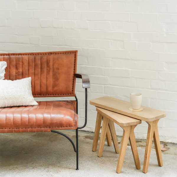 A set of solid wood Nested Side Tables made from solid Oak sits alongside a comfy leather sofa with a steaming cup of tea resting upon the larger of the two.