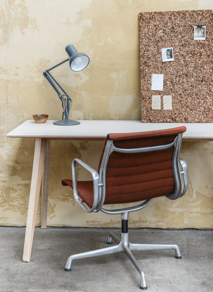 Brenin desk with light coloured lino top and oak veneer finish on the legs and underside with a table lamp on top, dark orange office desk tucked under and a cork board with notes attached leaning against the wall
