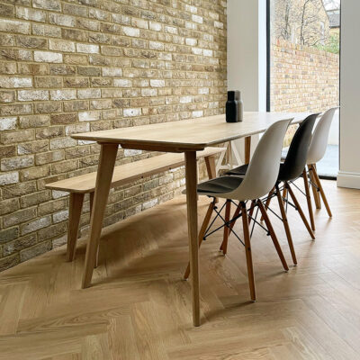 An elegant, solid oak wood Highbeach Dining Table and bench sit against an exposed brick wall and on a parquet wood flooring.
